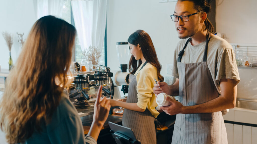 Young Asia male barista waiter taking order from customer standing behind bar counter while talking with customer making note on takeaway coffee cup at cafe restaurant. Owner small business concept.